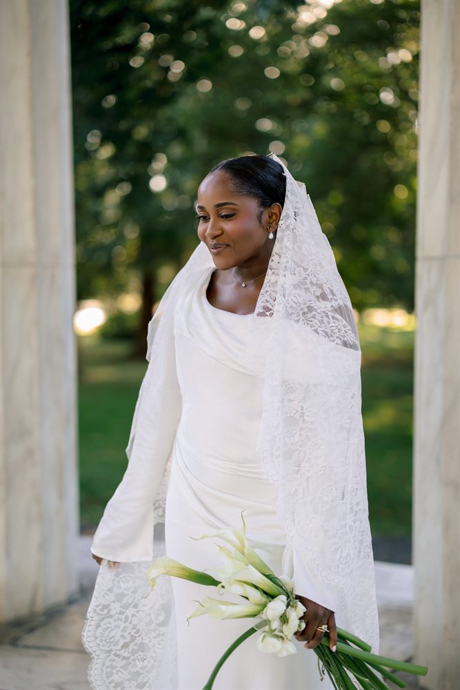 A bride in a lace mantilla veil holding calla lilies between the marble columns of the DC War Memorial
